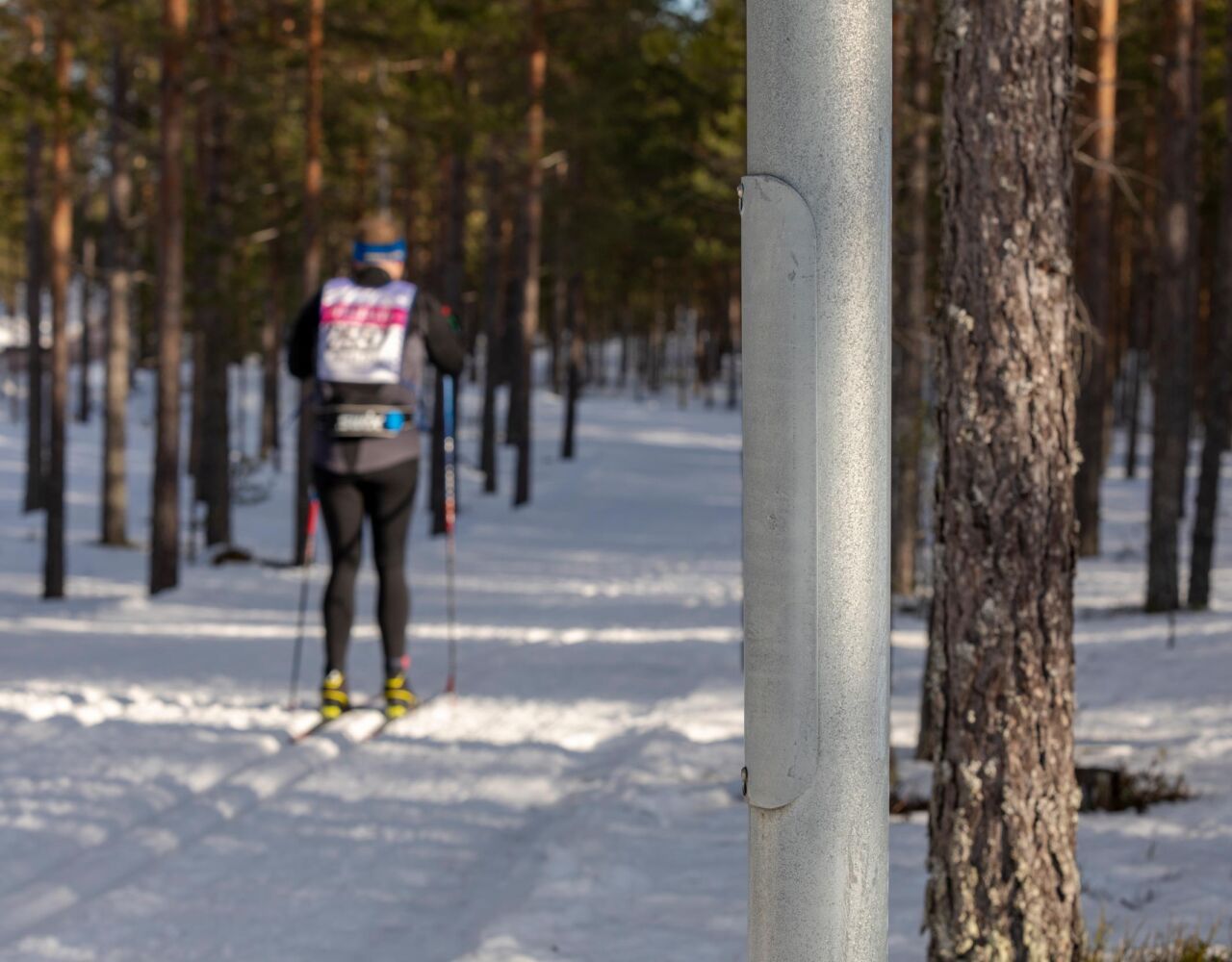 Skidåkare i Vasaloppsspåret med belysningsstolpe från Moramast i närbild i förgrunden. Foto: Kola Productions.>