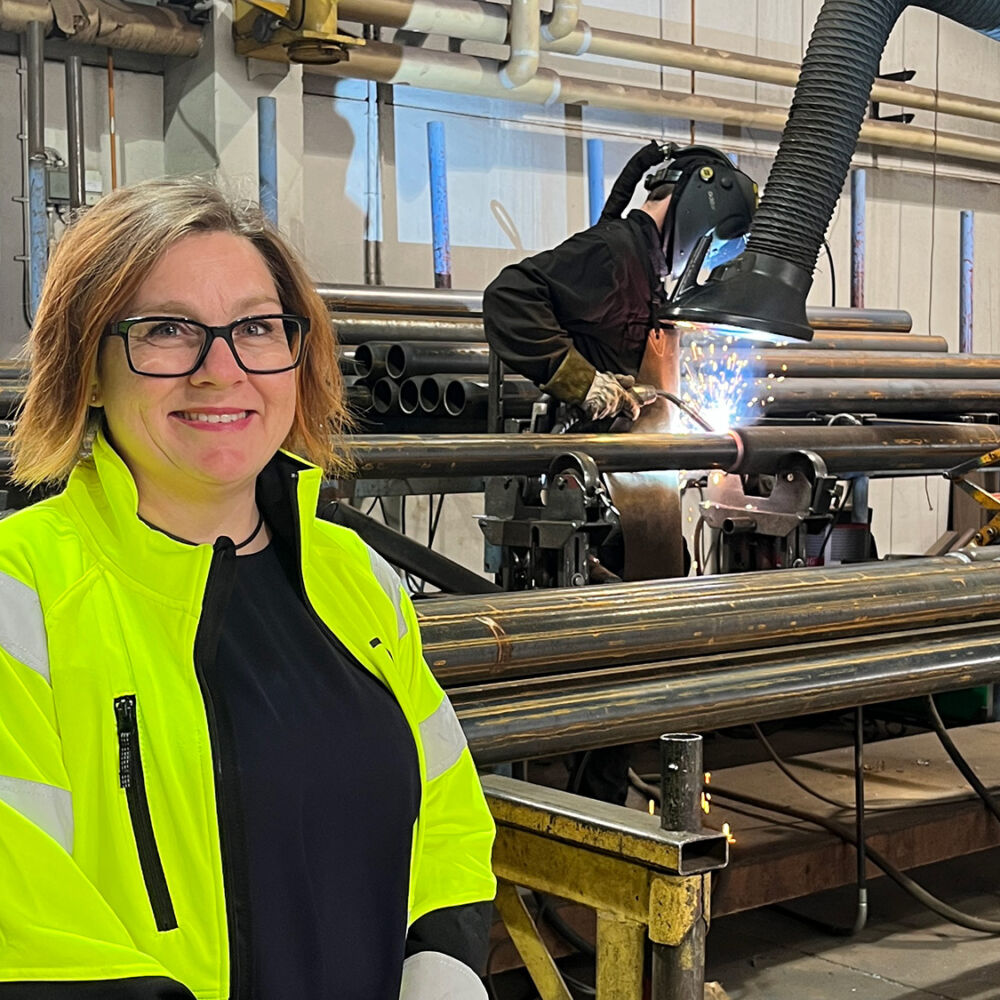 Ellinor Sparby, ny hållbarhetschef på Moramast, står i en av deras produktionslokaler med en svetsare i bakgrunden.