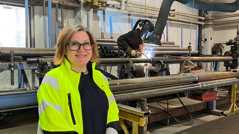 Ellinor Sparby, ny hållbarhetschef på Moramast, står i en av deras produktionslokaler med en svetsare i bakgrunden.