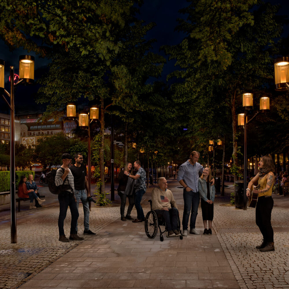 En trubadur spelar vid en lyktstolpe från Moramast i Kungsträdgården, Stockholm, medan folk lyssnar och dansar.